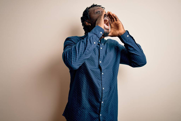 Mladý pohledný africký Američan muž na sobě ležérní košili stojící nad bílým pozadím křičí rozzlobený nahlas s rukama přes ústa - Fotografie, Obrázek