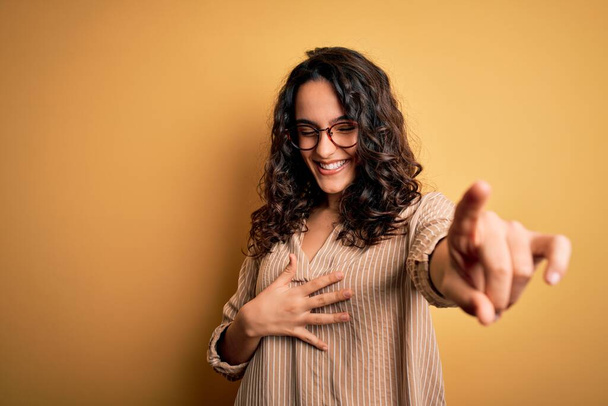 Beautiful woman with curly hair wearing striped shirt and glasses over yellow background laughing at you, pointing finger to the camera with hand over body, shame expression - Foto, Bild