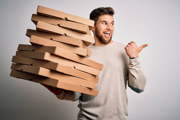 Young blond man with beard and blue eyes holding cardboards of pizza over white background pointing and showing with thumb up to the side with happy face smiling - Photo, Image
