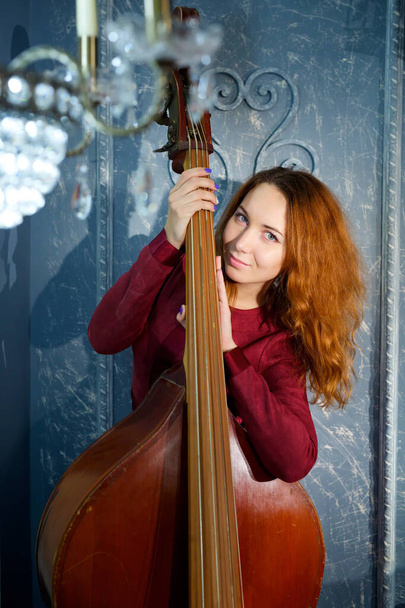 Dziewczyna w czerwonej sukience stojąca w studio trzymając instrument muzyczny kontrabas. Koncepcja muzyki klasycznej i jazzowej oraz wykonawstwa muzycznego. - Zdjęcie, obraz