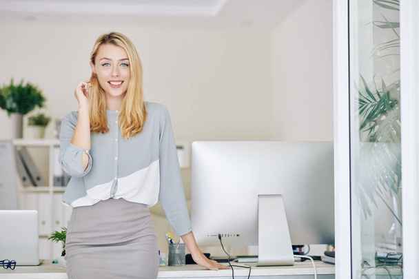 Piękny uśmiech młoda kobieta przedsiębiorca opierając się na stole biurowym i patrząc w aparat - Zdjęcie, obraz