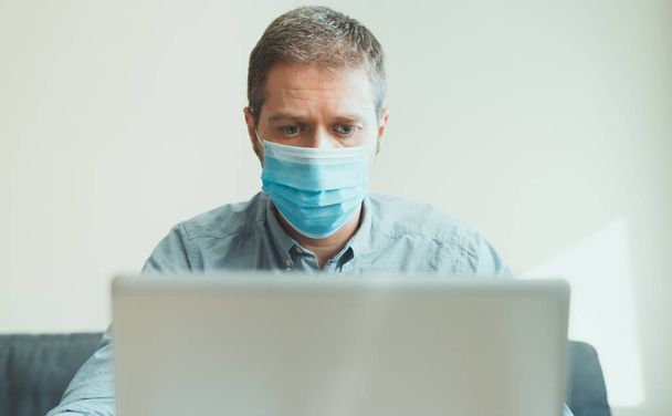 Άντρας με ιατρική μάσκα που εργάζεται στον υπολογιστή σε εσωτερικούς χώρους. - Φωτογραφία, εικόνα