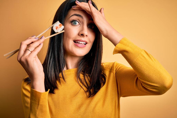 Mulher morena jovem com olhos azuis comendo sushi de maki de salmão usando pauzinhos estressados com a mão na cabeça, chocados com vergonha e rosto surpresa, irritados e frustrados. Medo e chateado por erro
. - Foto, Imagem