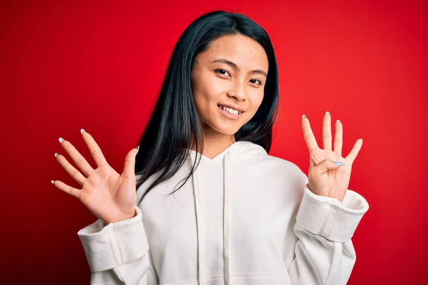 Joven hermosa mujer deportiva china con sudadera sobre fondo rojo aislado que muestra y señala hacia arriba con los dedos número nueve mientras sonríe confiado y feliz
. - Foto, imagen