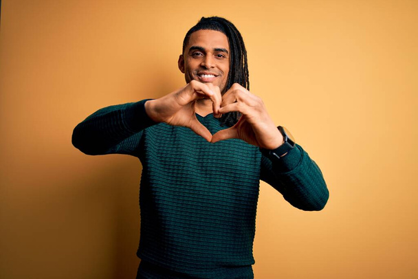 Fiatal, jóképű afro-amerikai afro férfi rasztával, zöld alkalmi pulóvert visel, szerelmes mosollyal, szív szimbólum alakot öltve a kezével. Romantikus koncepció. - Fotó, kép