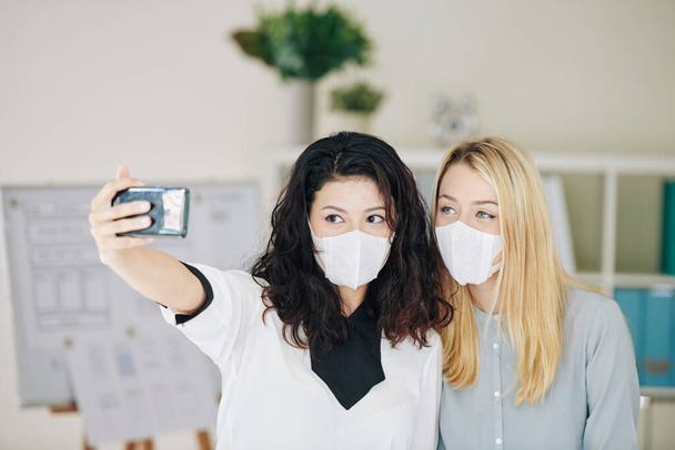 Junge Geschäftsfrauen in Arztmasken posieren im Büro für Selfie - Foto, Bild