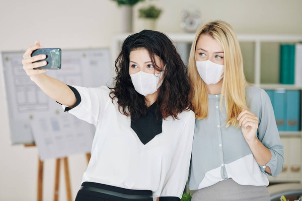 Hübsche junge Geschäftsfrau in medizinischer Maske arbeitet während der Pandemiezeit im Büro und macht Selfie mit Kollegin - Foto, Bild
