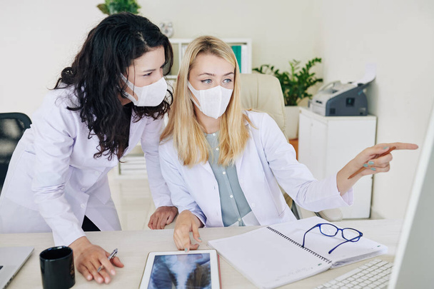 Young serious infectious disease specialist pointing at computer screen and showing results of medical test to coworker - Photo, Image