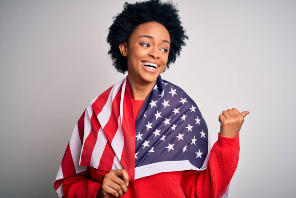 Jonge Afro-Amerikaanse afro vrouw met krullend haar in de Verenigde Staten van Amerika vlag wijzen en tonen met duim naar de zijkant met gelukkig gezicht glimlachend - Foto, afbeelding