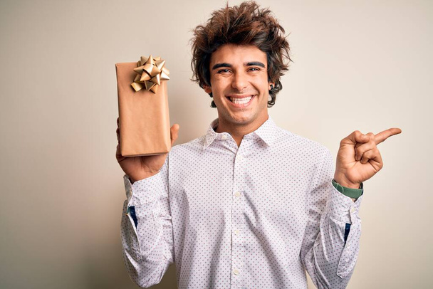 Молодий красивий чоловік тримає подарунок на день народження, стоячи над ізольованим білим тлом дуже щасливий, вказуючи рукою і пальцем на бік
 - Фото, зображення