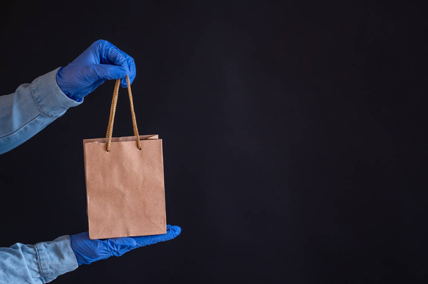 Female hands in blue gloves holds a brown paper bag with handles on a black background. Safe food delivery to your home. A courier in a denim shirt holds out a craft cardboard bag to a customer. - Photo, image