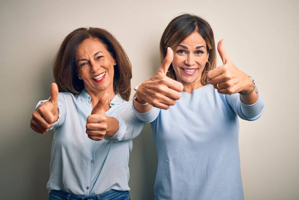 Middle age beautiful couple of sisters standing over isolated white background approving doing positive gesture with hand, thumbs up smiling and happy for success. Winner gesture. - Photo, Image