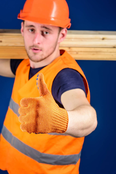 Carpenter, woodworker, builder on strict face carries wooden beam on shoulder. Wooden materials concept. Man in helmet and protective glove shows thumb up gesture, blue background, selective focus - Zdjęcie, obraz