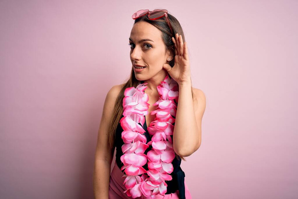 Fiatal, gyönyörű szőke nő fürdőruhát és virágos hawaii lejt visel rózsaszín háttér felett mosolyogva, fültől fülig hallgatva egy pletykát vagy pletykát. Süketség fogalma. - Fotó, kép