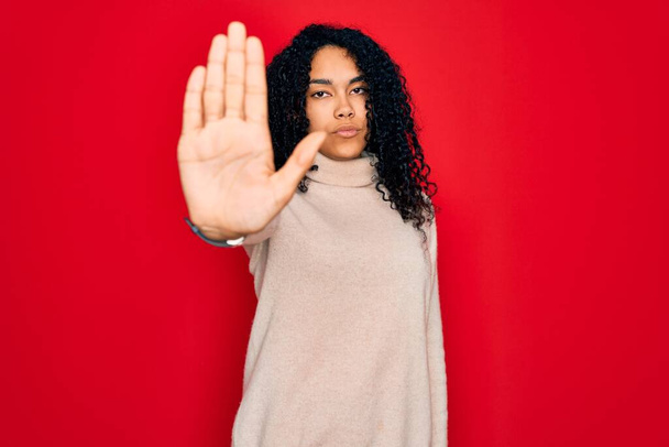Young african american curly woman wearing casual turtleneck sweater over red background doing stop sing with palm of the hand. Warning expression with negative and serious gesture on the face. - Photo, Image