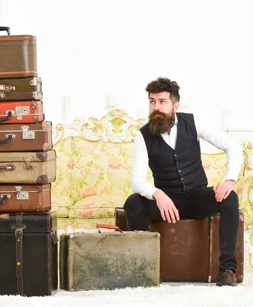 Man with beard and mustache packed luggage, white interior background. Luggage and relocation concept. Macho elegant on tired face sits, exhausted at end of packing, near pile of vintage suitcases - Фото, зображення