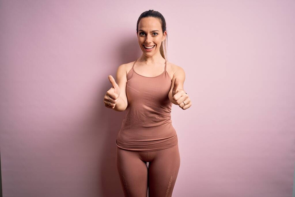 Young blonde fitness woman wearing sport workout clothes over pink isolated background approving doing positive gesture with hand, thumbs up smiling and happy for success. Winner gesture. - Photo, Image