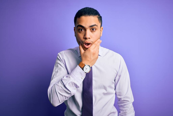 Молодой бразильский бизнесмен, одетый в элегантный галстук, стоящий на изолированном фиолетовом фоне, выглядит очарованным неверием, удивлением и изумленным выражением лица с руками на подбородке
 - Фото, изображение