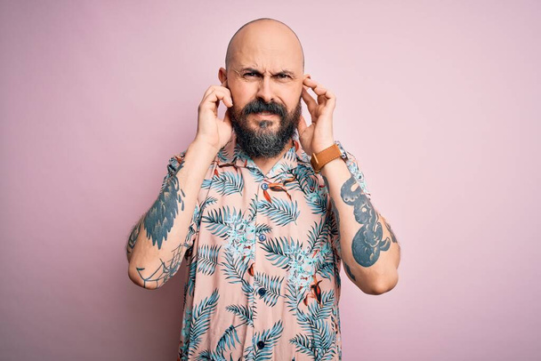 Hombre calvo guapo con barba y tatuaje con camisa floral casual sobre fondo rosa que cubre las orejas con dedos con expresión molesta por el ruido de la música fuerte. Concepto sordo
. - Foto, imagen