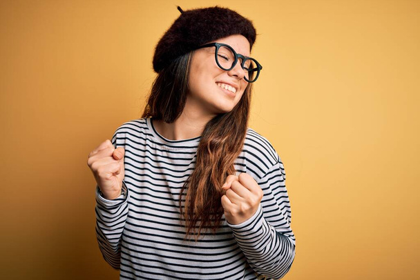 Młoda piękna brunetka nosi francuski beret i okulary na żółtym tle bardzo szczęśliwy i podekscytowany robi gest zwycięzca z podniesionymi ramionami, uśmiech i krzyk o sukces. Koncepcja uroczystości. - Zdjęcie, obraz