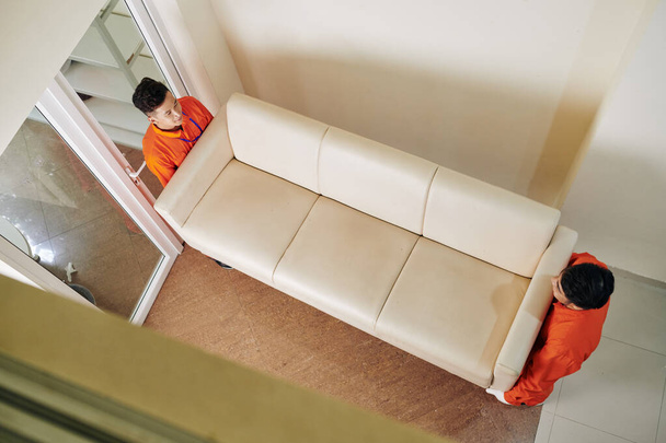Movers carrying heavy beige leather sofa in new house, view from above - Photo, Image