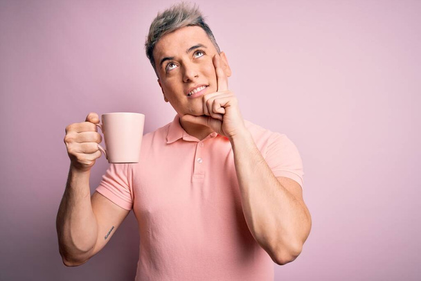 Young modern handsome man drinking a cup of hot coffee over isolated pink background serious face thinking about question, very confused idea - Zdjęcie, obraz