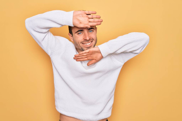 Young handsome man with blue eyes wearing casual sweater standing over yellow background Smiling cheerful playing peek a boo with hands showing face. Surprised and exited - Photo, Image