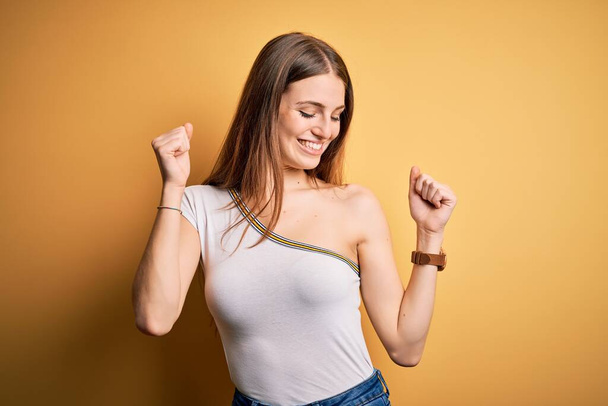 Junge schöne rothaarige Frau trägt lässiges T-Shirt über isoliertem gelben Hintergrund Tanzen glücklich und fröhlich, lächelnd bewegt sich lässig und selbstbewusst Musik hören - Foto, Bild