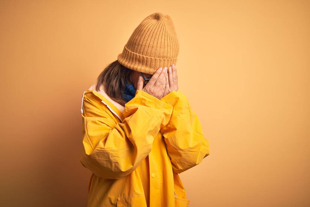 Sarı yağmurluk ve kış şapkası takan orta yaşlı bir kadın izole bir arka planda üzgün bir ifadeyle elleri ile yüzünü kapatırken ağlıyordu. Depresyon kavramı. - Fotoğraf, Görsel