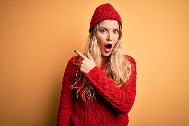 Mladá krásná blondýnka žena na sobě ležérní svetr a vlněná čepice přes bílé pozadí Překvapený ukazující prstem na stranu, otevřená ústa užaslý výraz. - Fotografie, Obrázek