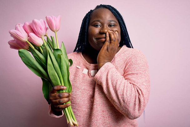 Junge afrikanisch-amerikanische Plus-Size-Frau mit Zöpfen, die einen Strauß rosa Tulpenblüten in der Hand hält, lacht und verlegen kichert, den Mund mit Händen, Klatsch und Skandalkonzept bedeckt - Foto, Bild