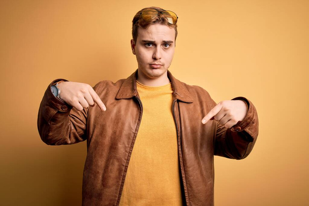 Young handsome redhead man wearing casual leather jacket over isolated yellow background Pointing down looking sad and upset, indicating direction with fingers, unhappy and depressed. - Photo, Image