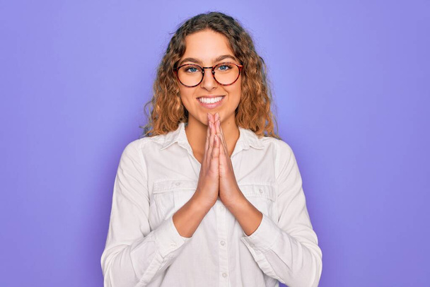 Young beautiful woman with blue eyes wearing casual shirt and glasses over purple background praying with hands together asking for forgiveness smiling confident. - Fotó, kép