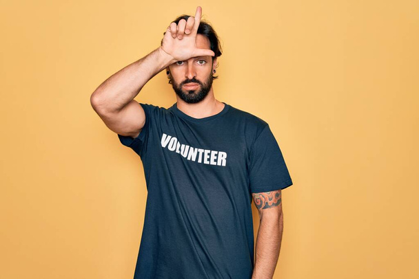 Fiatal jóképű spanyol önkéntes férfi önkéntes pólót visel szociális gondozás gúnyolódik az emberek ujjak a homlokán csinál vesztes gesztus gúnyolódik és sértő. - Fotó, kép