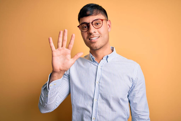 Jovem homem de negócios hispânico bonito usando óculos nerd sobre fundo amarelo mostrando e apontando para cima com os dedos número cinco enquanto sorri confiante e feliz
. - Foto, Imagem