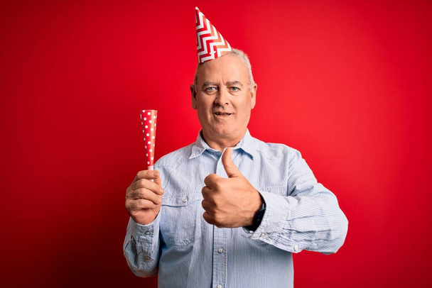 Homem de meia idade usando chapéu engraçado usando trompete celebrando aniversário sobre fundo vermelho feliz com grande sorriso fazendo sinal ok, polegar com os dedos, excelente sinal
 - Foto, Imagem