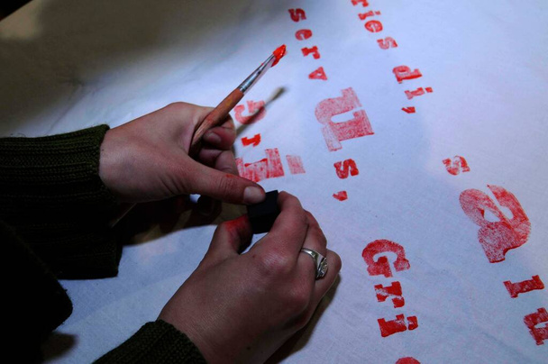 punaisten kirjainten painaminen valkoiselle tekstiilille käsin ja harjalla
 - Valokuva, kuva