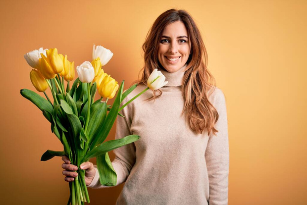 Junge schöne brünette Frau mit einem Strauß gelber Tulpen vor isoliertem Hintergrund und einem fröhlichen und kühlen Lächeln im Gesicht. Glücklicher Mensch. - Foto, Bild