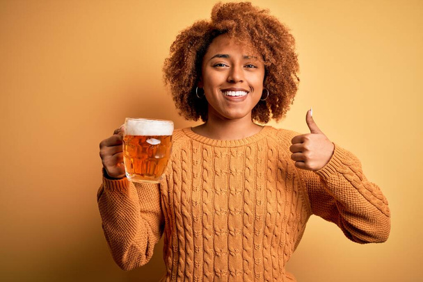 Jonge mooie Afro-Amerikaanse afro vrouw met krullend haar drinken pot bier blij met grote glimlach doet ok teken, duim omhoog met vingers, uitstekend teken - Foto, afbeelding