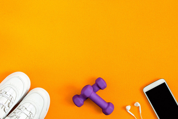 Sneakers, halters, smartphone met hoofdtelefoon op achtergrond van oranje fitnessmat. Vlakke lay compositie. Concept van sport, fitness, gezonde levensstijl. Bovenaanzicht. Kopieerruimte. - Foto, afbeelding