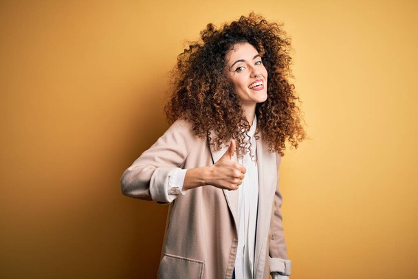 Young beautiful brunette woman with curly hair and piercing wearing casual t-shirt and diadem doing happy thumbs up gesture with hand. Approving expression looking at the camera showing success. - Photo, Image