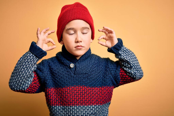 Молодий маленький хлопчик, який носить вовняну шапку і зимовий светр над жовтим ізольованим тлом, розслабляється і посміхається очима, закритими роблячи медитаційний жест пальцями. концепція йоги
. - Фото, зображення