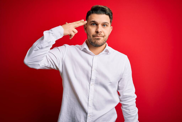 Young business man with blue eyes wearing elegant shirt standing over red isolated background Shooting and killing oneself pointing hand and fingers to head like gun, suicide gesture. - Photo, Image
