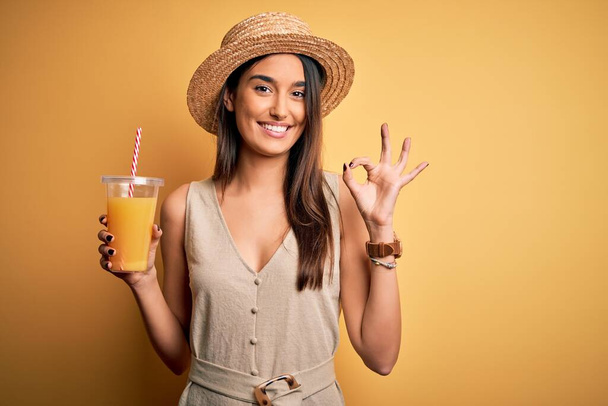 Młoda piękna kobieta na wakacjach w letniej czapce pije zdrowy sok pomarańczowy robi ok znak palcami, doskonały symbol - Zdjęcie, obraz