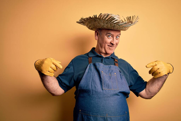 Homem agricultor hoary meia idade vestindo avental e chapéu sobre fundo amarelo isolado olhando confiante com sorriso no rosto, apontando-se com os dedos orgulhosos e felizes
. - Foto, Imagem