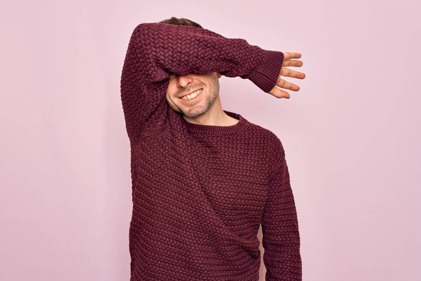 Jonge knappe man met blauwe ogen met casual trui over roze achtergrond die ogen bedekt met een arm die vrolijk en grappig glimlacht. Blind concept. - Foto, afbeelding