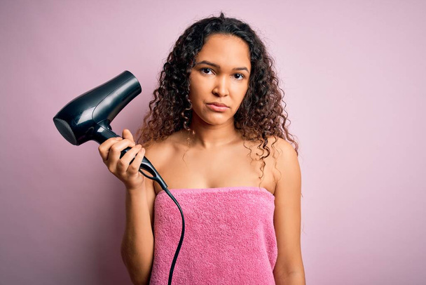 Beautiful woman with curly hair wearing shower towel after bath using hair dryer with a confident expression on smart face thinking serious - Photo, Image