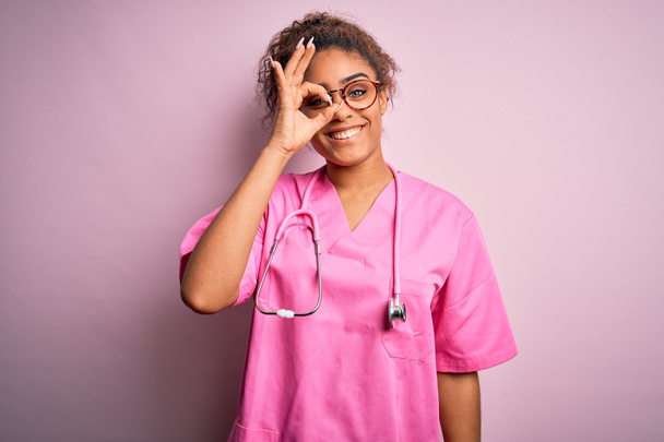 Африканская американская медсестра девушка в медицинской форме и стетоскоп на розовом фоне делает хорошо жест с улыбкой на руке, глаза смотрят сквозь пальцы с счастливым лицом
. - Фото, изображение