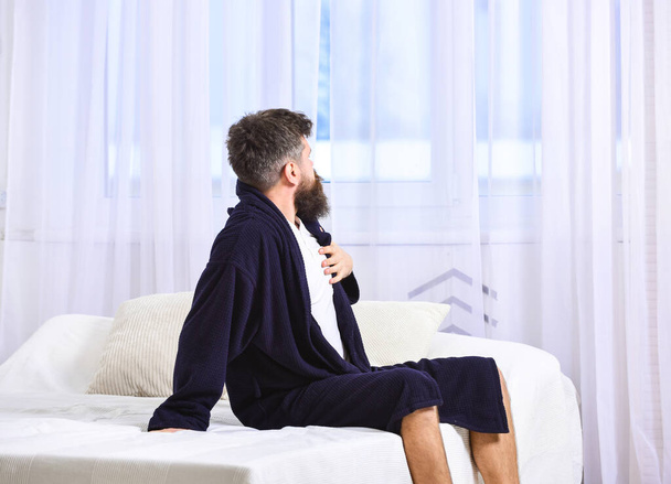 Guy sleepy looking at window in morning. Awakening concept. Macho with beard and mustache sluggish relaxing after nap, rest. Man in robe sits on bed, white curtains on background. - 写真・画像