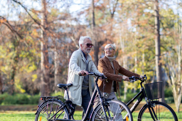 Mature fit couple pushing bicycles in public park talk and smile - Foto, Imagem
